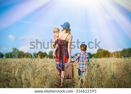Picture of woman wearing hat with baby girl and boy walking away on wheat field. She carrying child and big old suitcase over blue sky sunny outdoors background