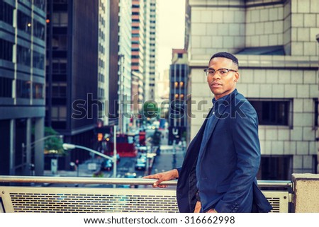 African American college student studying in New York. Wearing blue jacket, glasses, a young black man standing by railing on balcony, facing street, thinking, lost in thought. Instagram effect.

