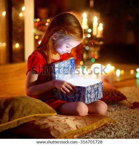 Adorable little girl opening a magical Christmas gift by a Christmas tree in cozy living room in winter