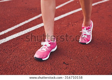 Young girl legs in pink sneakers front side on a red race track of a stadium with white lane lines on background. Unrecognizable photo with copy space