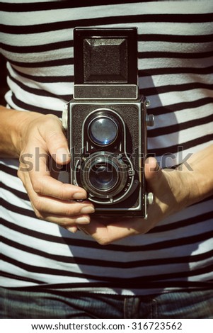 Middle aged woman in striped shirt and jeans is holding an old camera in her hands and is ready to take pictures. Photo is edited as a vintage photo. All potential trademarks are removed.