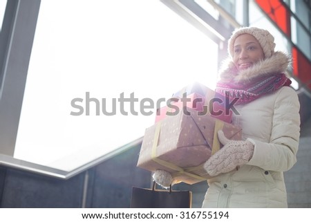 Smiling woman in warm clothing carrying stacked gifts by window