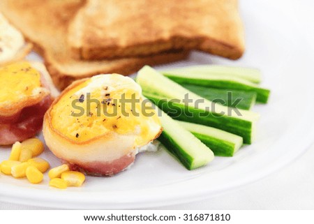 Baked eggs with bacon, cucumber, corn and toast on a plate, white background.