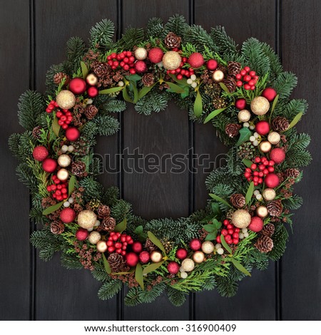 Christmas wreath with red and gold bauble decorations, bow, holly, mistletoe, pine cones and blue spruce fir over dark oak front door background.