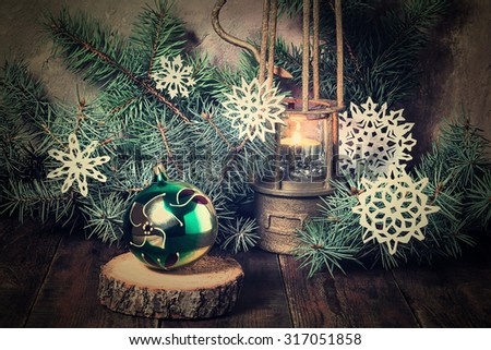Old bronze lamp with a burning candle in the branches of a blue Christmas tree, decorated with paper snowflakes, Christmas ball on a wooden stand. Tinted photos