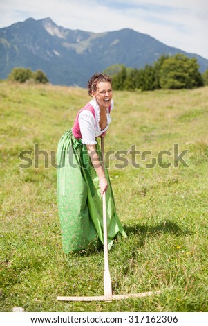 Peasant woman in middle age in dirndl having fun while working on a mountain meadow with a rake