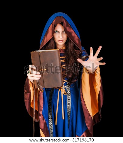 Beautiful young halloween witch wearing vintage gothic dress with hood holding magical book of spells in old leather cover making magic isolated on black background