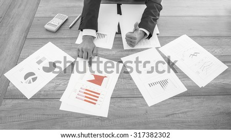 Greyscale image with selective color of a successful businessman pointing to a red chart amongst four spread out on his desk giving a thumbs up of approval, high angle view of the desk and his hands.