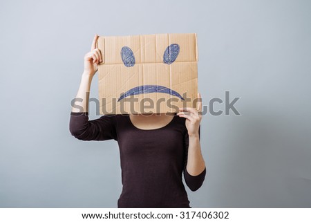 A woman holds a sad smiley on cardboard. On a gray background.