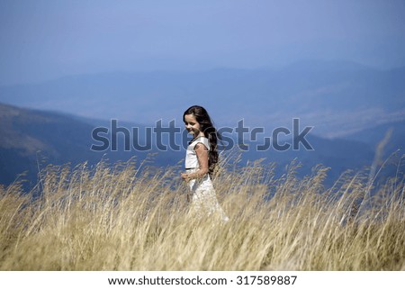 Pretty smiling brunette girl in white lace summer dress walking in mountain valley among deep dry spikelet grass in spring sunny day outdoor on natural blue background, horizontal picture