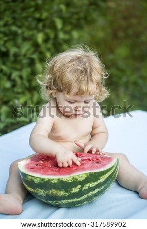 One little boy with blond curly hair and round cheeks sitting on blue plaid holding half of fresh juicy green and red water melon with black seeds looking down on natural background, vertical picture