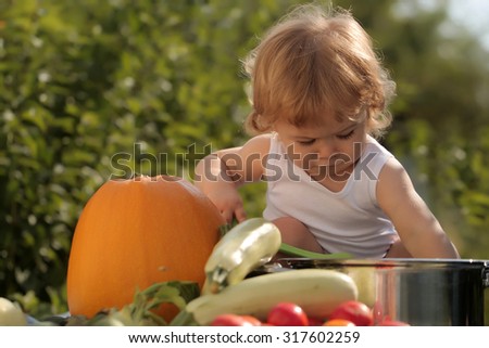 Cute small kid at picnic holding ladle sitting near pot among orange pumpkin red tomato squash and cucumber on blue checkered plaid on natural background sunny day, horizontal picture