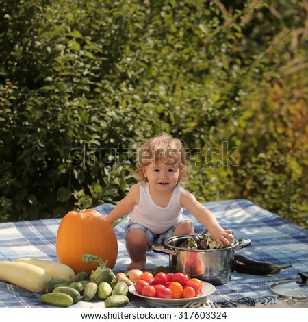 One little playful boy at picnic playing and sitting near pot orange pumpkin squash and cucumber red tomato on checkered plaid looking forward on natural background sunny day, square picture