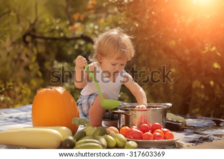 One little baby boy at picnic sitting and playing with ladle pot orange pumpkin squash and cucumber red tomato on checkered plaid looking forward on natural background sunny day, horizontal picture