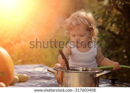 Small boy at picnic holding ladle sitting near pot among orange pumpkin red tomato squash and cucumber playing with food on blue checkered plaid on natural background sunny day, horizontal picture