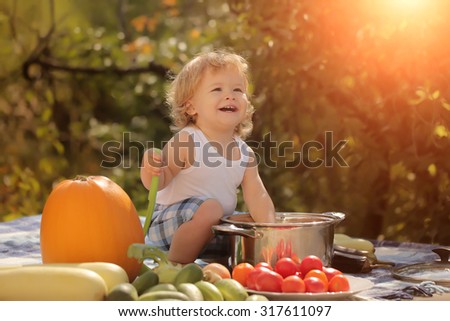 One laughing boy at picnic sitting with ladle pot orange pumpkin red tomato squash and cucumber playing with food sitting on blue checkered plaid on natural background sunny day, horizontal picture