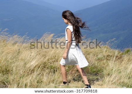 Back view of young little brunette girl in white lace summer dress walking in mountain valley with deep dry spikelet grass in spring sunny day outdoor on natural blue background, horizontal picture