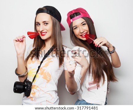  Fashion portrait of two young pretty hipster girls wearing bright make up and holding candys. Studio portrait of two cheerful best friends sisters having fun and making funny faces.