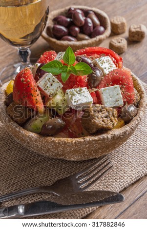 At the center of vertical frame the bread bowl of Greek salad on a table-napkin of burlap in the background olives, glass of wine, croutons.  Cretan Greek salad for lunch, snacks. Vertical shot. 


