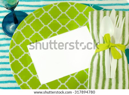 A casual place setting in green and turquoise with fun shapes on plates and white plastic utensils. Striped towel background. Cloth napkin tied with green ribbon. Card blank for text. Overhead view.