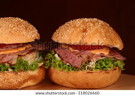 Two appetizing big delicious fresh burgers with green lettuce red tomato cheese cabbage bacon slice meat cutlet and white bread bun with sesame seeds on black background closeup, horizontal picture