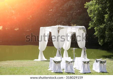 Beautiful decorated with light white chiffon chandelier and bouquets of roses pastel colors wedding pavilion chairs and table standing on green grass near lake and trees sunny day, horizontal picture