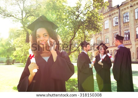 Young students dressed in black graduation gown. Campus as a background. Afro American girl cheerfully smiling, holding diploma and talking on phone. Students are on background. Light flare on photo