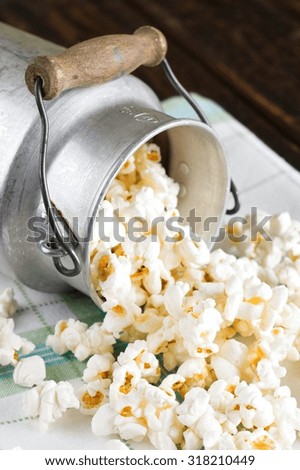 Vertical photo with old aluminum can which is placed on green towel and old wooden board which is full of popcorn spilled out on the cloth.