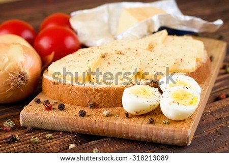 Horizontal photo with halves of quail eggs on chopping board with bread with butter and slices of romadur ripened cheese and tomatoes with onion around on wooden board with whole pepper.