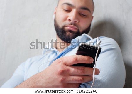 In the rhythm of my music. Relaxed young bearded man in headphones with smartphone leaning on the grey wall and listening to the music. Focus on phone.