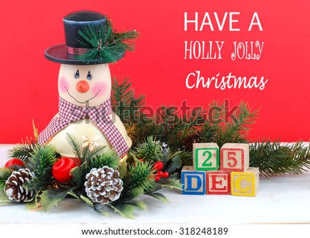 A cute snowman decoration with lamp inside is surrounded by holly, pine, pine cones and Christmas decorations. Bright red background. Have a Holly Jolly Christmas message with Dec. 25 in letter blocks