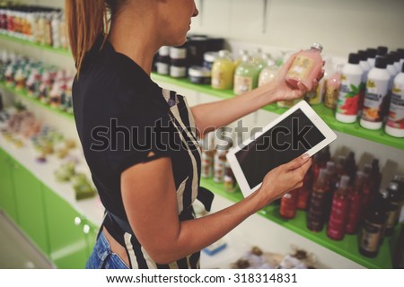 Cropped image of saleslady holding cosmetic product in plastic bottle with blank space label while using digital tablet with copy space screen, woman seller reads contents information of the product