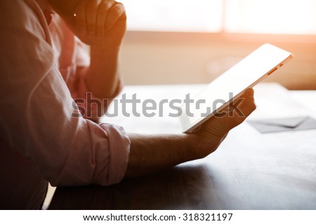 Thoughtful male person looking to the digital tablet screen while sitting in modern loft interior at the table, experienced entrepreneur reading some text or electronic book at the office, filter sun Royalty-Free Stock Photo #318321197
