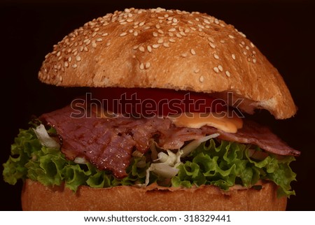 One appetizing big delicious fresh burger with green lettuce red tomato cheese and bacon slice and white bread bun with sesame seeds on black background closeup, horizontal picture