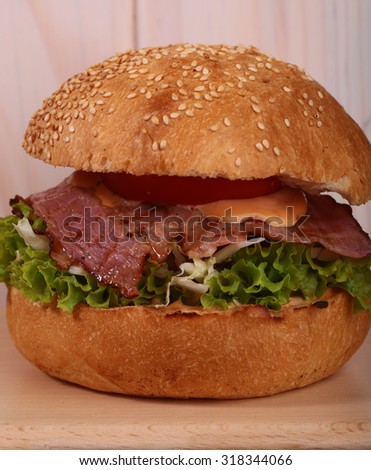 One appetizing big delicious fresh burger with green lettuce red tomato cheese and bacon slice and white bread bun with sesame seeds on wooden background closeup, square picture