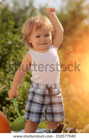 Smiling little curious boy in checkered summer shorts at picnic with ladle standing outdoor on green natural background sunny day, vertical picture