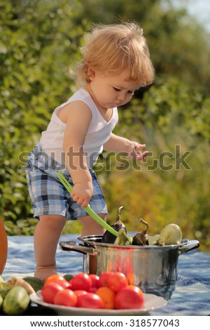 One little curious boy in shorts at picnic with ladle standing near pot red tomato orange pumpkin squash and cucumber on checkered plaid on natural background sunny day, vertical picture