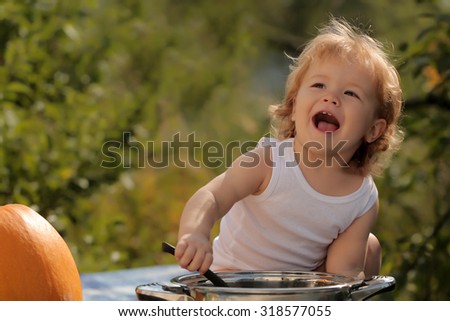 One happy smiling small child boy with blonde curly hair making food in kitchen bowl sitting at picnic with orange pumpkin sunny day outdoor on green natural background, horizontal picture