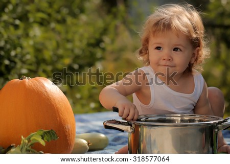 One curious playful little child boy with blonde curly hair making food in kitchen bowl sitting at picnic with orange pumpkin sunny day outdoor on green natural background, horizontal picture