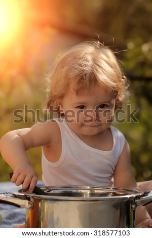 Portrait of smiling baby boy with blonde curly hair in white underwear holding kitchen utensil and cooking in big pot looking forward outdoor sunny day on green natural background, vertical picture