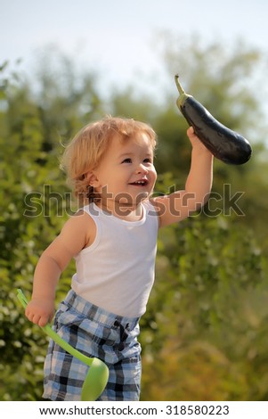 Happy little smiling boy in checkered summer shorts at picnic with ladle and purple eggplant standing outdoor on green natural background sunny day, vertical picture