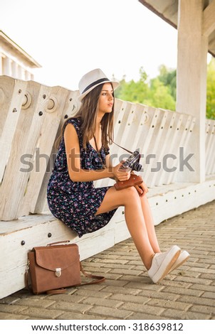 Side view of young woman in dress and hat is sitting outdoors with old camera.