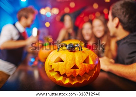 Carved pumpkin and spider on background of group of friends celebrating Halloween in bar