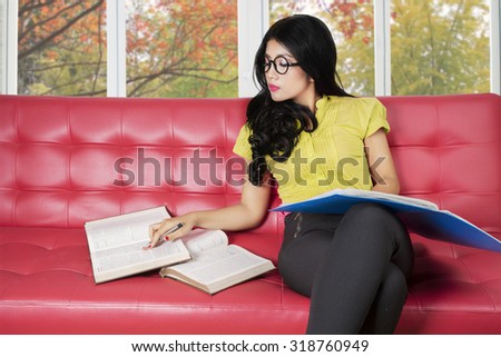 Photo of pretty college student sitting on the sofa while reading textbooks with autumn background on the window