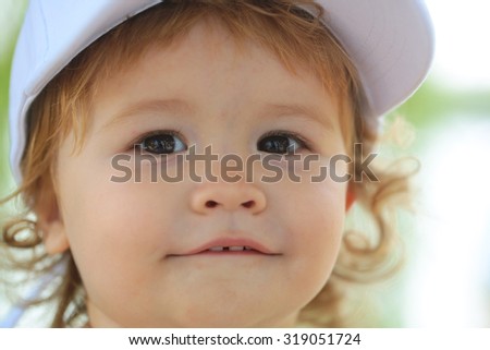 Portrait of one small happy smiling male kid swith blond curly hair in white cap on green natural blur background closeup, horizontal picture