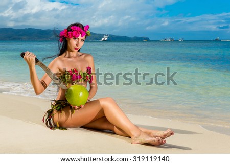 Young beautiful girl in a bikini made of flowers sitting on a tropical beach and coconut with a machete. Blue sea in the background.