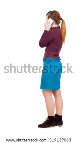 back view of standing young beautiful  woman and using a mobile phone. girl  watching. Rear view people collection. girl in a blue skirt and jacket of red and white talking cell phone.