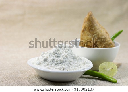 Finely milled refined wheat flour (maida) in a white bawl; a samosa (A triangular savory pastry fried in oil) made from this wheat flour served with two green chilies and a lemon cut in half. Royalty-Free Stock Photo #319302137