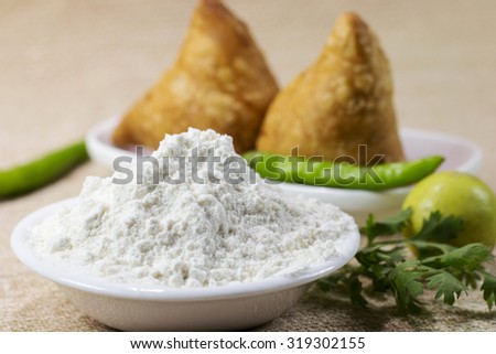 Finely milled refined wheat flour (maida) in a white bawl ; two samosas made from this wheat flour served with two green chilies, a lemon and some coriander leaves (in the rear and out of focus). Royalty-Free Stock Photo #319302155