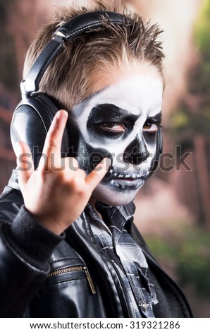 Close up portrait of young tough rocker boy with skull make up wearing music headphones and black leather jacket and holding rock on sign